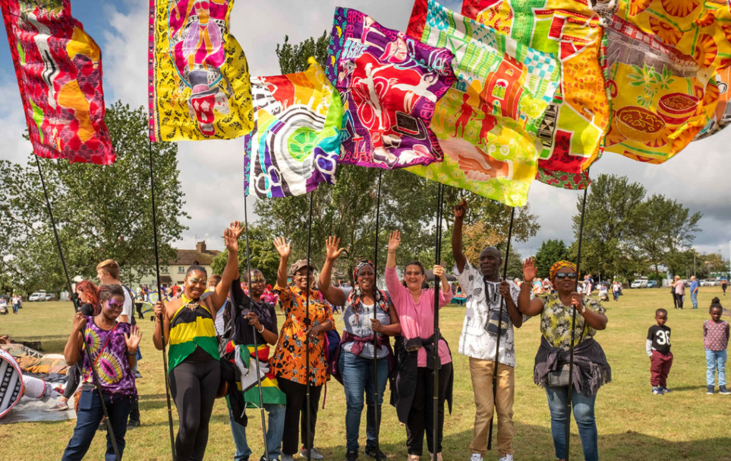 Large colourful flags are held by a group of smiling people waving at the camera